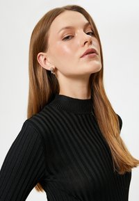 Schwarzer gerippter Pullover mit vertikalen Streifen und hohem Kragen. Das Model hat lange, glatte Haare und trägt schlichte Ohrringe.