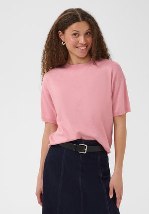 Femme aux cheveux bouclés souriante, portant un haut rose à manches courtes rentré dans une jupe en jean foncé avec une ceinture noire sur un fond uni.
