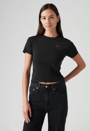 Femme aux longs cheveux foncés portant un t-shirt court noir Levi's et un jean noir taille haute, debout, une main sur la hanche, sur un fond uni.