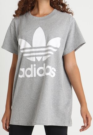Kobieta w luźnej szarej koszulce z dużym białym logo Adidas i napisem z przodu, stojąca na tle jednolitego tła.