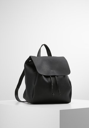 Sac à dos en cuir noir texturé avec fermeture à rabat et cordon, posé sur un piédestal blanc devant un fond clair uni.