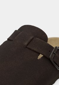 Καφέ σανδάλι από σουέντ με φαρδιά λουράκι και ρυθμιζόμενο μεταλλικό πόρπη. Διαθέτει ελαφριά ανάγλυφη σόλα και μινιμαλιστικό, ανοιχτό σχέδιο.