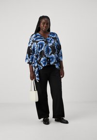 Svart blomstret bluse med blå blomsterdesign, V-hals, løs passform, kombinert med svarte bukser og en liten hvit veske. Svarte sko fullfører antrekket.