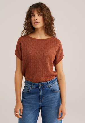 Joven con cabello rizado vistiendo una blusa de punto con estampado color óxido y jeans azules de tiro alto, frente a un fondo liso.