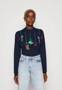 Marineblå genser med festlige paljettmønstre, inkludert et juletre, sukkertøystokker, hjerter og et reinsdyr, kombinert med lyse jeans.