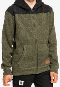 Sudadera verde con cremallera y panel superior negro texturizado, que presenta bolsillos frontales y un parche de cuero en el dobladillo inferior.