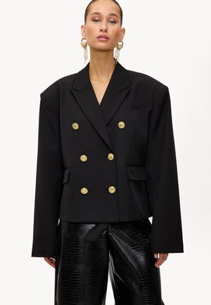 Mujer vistiendo una chaqueta negra de gran tamaño, de corte cruzado, con botones dorados y pantalones de tiro alto texturizados en negro, acompañada de grandes pendientes de aro dorados.