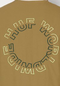 Brązowy bawełniany t-shirt z okrągłym, haftowanym napisem w zielonym i jasnżółtym kolorze, z napisem "HUF WORLDWIDE" na plecach.