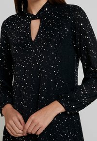 Černé šaty s dlouhými rukávy, výřezem na krku a stříbrnými tečkovanými ozdobami. Texturovaná tkanina dodává designu hloubku.
