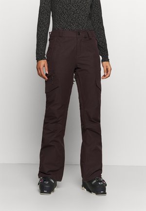 Person trägt schwarzen gemusterten Langarmtop, dunkelbraune Cargohosen und schwarze Skistiefel, steht auf hellem Boden vor einer weißen Wand.