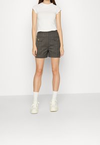 Pantaloni corti grigi a quadri con tasche frontali e dettagli in bottone oro, abbinati a una T-shirt bianca e sneakers bianche, con polsini a coste.