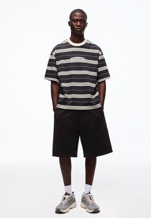 Férfi fekete-fehér csíkos pólóban, fekete térdig érő rövidnadrágban, fehér zokniban és szürke futócipőben, zsebében tartva a kezét áll.