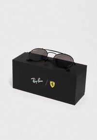 Στρογγυλά μαύρα γυαλιά ηλίου με σκουρόχρωμους φακούς, μεταλλικό σκελετό, τοποθετημένα σε μαύρο κουτί που φέρει τα λογότυπα της Ray-Ban και της Ferrari.