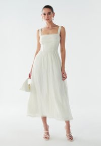 Balta midi kleita ar kvadrātveida izgriezumu, fakturētu audumu, mežģīņu apdari uz plecu siksnām un apakšmalu, kombinēta ar baltiem sandales ar siksniņām un somiņu.