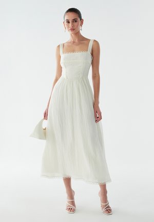 Balta midi suknelė kvadratinėmis iškirptėmis, su nėrinių apdaila ir lengva tekstūra. Modelis laiko baltą rankinę ir dėvi baltus šlepetes su dirželiais.