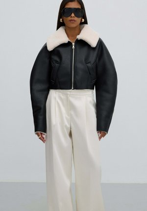 Zwarte leren cropped jack met een crèmekleurige faux-fur kraag, met een ritssluiting en brede, gestructureerde mouwen, gecombineerd met witte wijde broekspijpen.