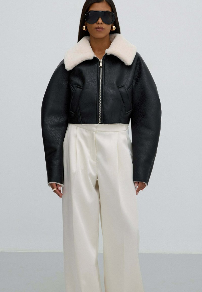 Zwarte leren cropped jack met een crèmekleurige faux-fur kraag, met een ritssluiting en brede, gestructureerde mouwen, gecombineerd met witte wijde broekspijpen.