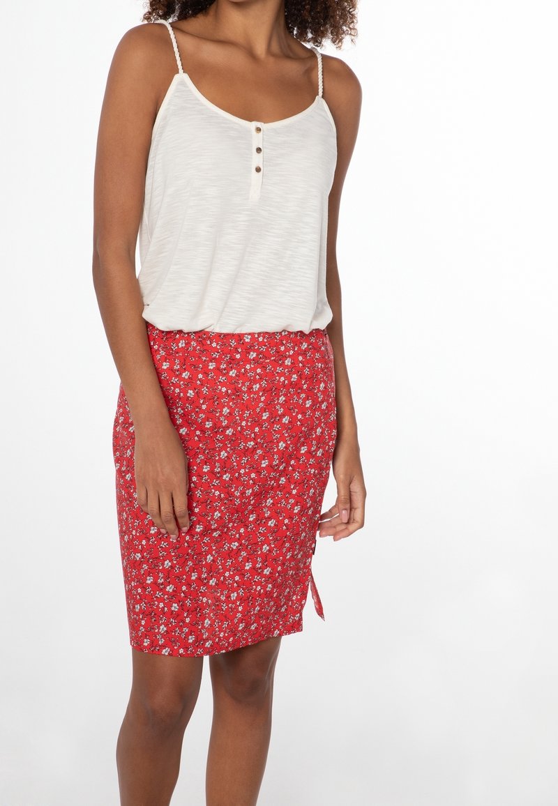 Femme portant un haut blanc sans manches boutonné et une jupe rouge fleurie longueur genou avec une petite fente latérale.
