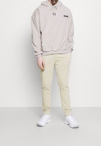 Beige fleece hoodie med justerbar dragsko, framficka och logotypdetalj, ihop med ljusbeiga joggerbyxor och vita sneakers.