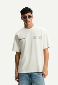 Crème-kleurig katoenen T-shirt met korte mouwen, met een zwarte print van luchthavencodes en 'Departures' aan de linkerkant. Gedragen met blauwe denim jeans.
