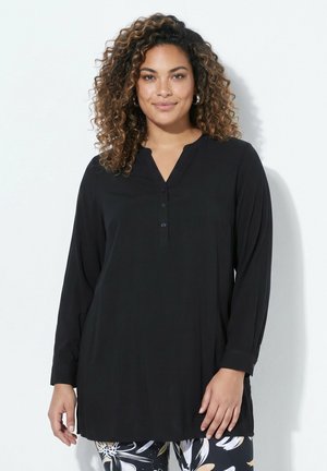 Frau mit lockigem Haar trägt ein langärmliges schwarzes Tunika-Oberteil und gemusterte schwarze, weiße und beige Hosen vor einem schlichten hellen Hintergrund.