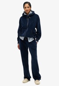 Chándal de terciopelo azul marino con chaqueta con capucha y cremallera y pantalones a juego, que presenta un corte relajado y detalles de cordón.