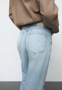 Svijetloplave traperice s visokim strukom, sa dva stražnja džepa i teksturiranim smeškim oversized puloverom. Vidljivo dekorativno šivanje.