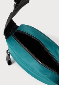 Τσαντάκι χρώματος τυρκουάζ, κατασκευασμένο από λείο υλικό, με μαύρο φερμουάρ και αποσπώμενο λουρί. Το εσωτερικό είναι σκοτεινό και χωρίς επένδυση.