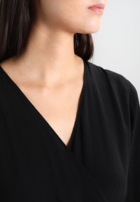 Sort indpakningsbluse med en dyb V-udskæring og blødt, glat stof. Krave-design med subtil tekstur, der fremhæver nakken og skuldrene.