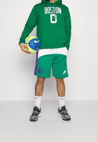 Felpa verde con il testo "BOSTON", pantaloni corti sportivi verdi e bianchi, scarpe da ginnastica nere e grigie, mentre tiene un pallone da basket multicolore.