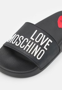 Sandale slide noire texturée avec une large bride affichant un texte blanc en relief "LOVE MOSCHINO" et un détail cœur rouge sur la semelle.