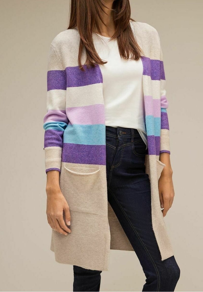 Femme portant un long cardigan beige avec de larges rayures horizontales pastel violet, rose et bleu, associé à un haut blanc et un jean foncé.