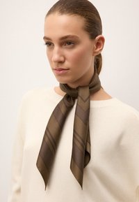 Brun silkeslørf med chevronmønster, knyttet om halsen, båret over en lys beige sweater. Tekstureret stof og elegant design.