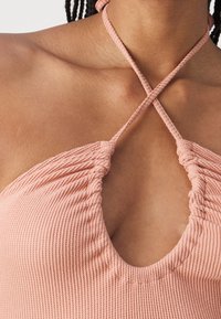 Haut halter en rose côtelé présentant un décolleté plongeant et des bretelles croisées au cou, avec une texture froncée au niveau de la poitrine et du haut du corps.