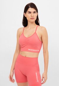 Sportiva color corallo con parte anteriore arricciata, spalline regolabili e logo "SQUATPROOF" in bianco. Abbinata a pantaloni corti a vita alta coordinati.