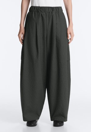 Osoba nosí široké čierne textúrované nohavice s elastickým pásom a šnúrkami, v kombinácii s čiernymi topánkami a bielym topom.