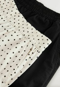 Deux pantalons à taille élastique, l'un entièrement noir et l'autre blanc à pois noirs, disposés à plat et superposés sur une surface blanche.