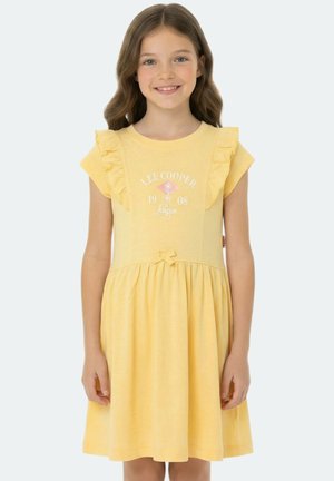 Jeune fille aux cheveux bruns ondulés portant une robe jaune à manches volantées avec le texte "Lee Cooper 1908 London" sur la poitrine.