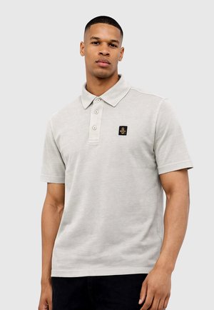 Giovane uomo con indosso una polo a maniche corte grigio chiaro con tre bottoni e un piccolo logo nero e oro sul petto.