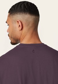 Korte paarse t-shirt gezien vanuit de achterkant, met een ronde halslijn en een klein geborduurd logo nabij de kraag. Gladde stof textuur.