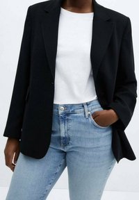 Blazer negro con un diseño ajustado, dos bolsillos frontales, llevado sobre una camiseta blanca y jeans azul claro. Tejido suave y forma estructurada.