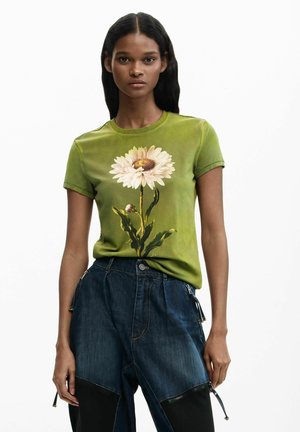 Young woman wearing green t-shirt with large white daisy print and dark blue pants with black patches and zippers.