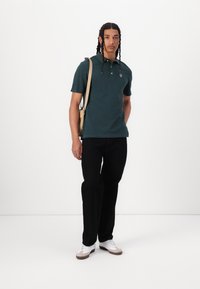 Mørkegrønn polo skjorte med krage, tre knapper og logo. Sorte bukser og hvite sneakers med grå detaljer. Brun skulderveske.