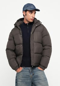 Brun oppustelig jakke med hætte, der har syede sektioner og knaplukninger, båret over en mørk sweater, sammen med lyse denim jeans og en blå kasket.