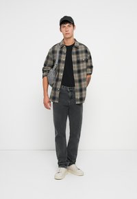 Camisa de flanela xadrez em verde e preto, t-shirt preta, jeans cinzentos, sapatilhas brancas, saco transversal cinzento e boné preto, em pé sobre um fundo branco.