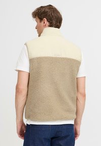 Beige Fleece-Weste mit einem glatten cremefarbenen Oberteil, einem hohen Kragen und einem strukturierten unteren Abschnitt. Auffälliges Markenetikett auf der Rückseite.