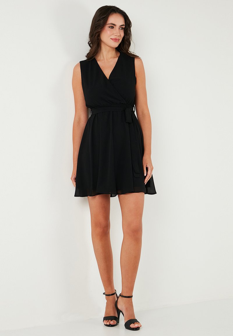 Zwarte mouwloze wikkeldress met een aansluitend lijfje en een uitlopende rok. Beschikt over een strikceintuur bij de taille en zachte, vloeiende stof.