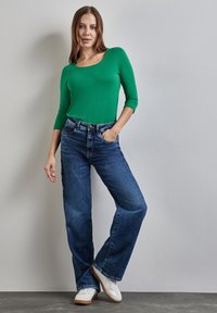 Vihreä pitkähihaine paita, jossa on pyöreä kaula-aukko, yhdistettynä sinisiin laidallisiin farkkuihin. Malli seisoo itsevarmana, kädet taskuissa, valkoiset tennarit jalassa.