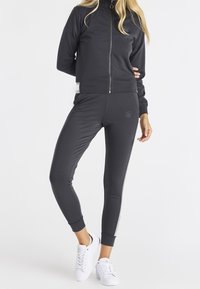 Survêtement gris foncé avec une veste zippée et un pantalon ajusté. La veste présente un col haut et un logo ; le pantalon a des rayures latérales et un détail logo.