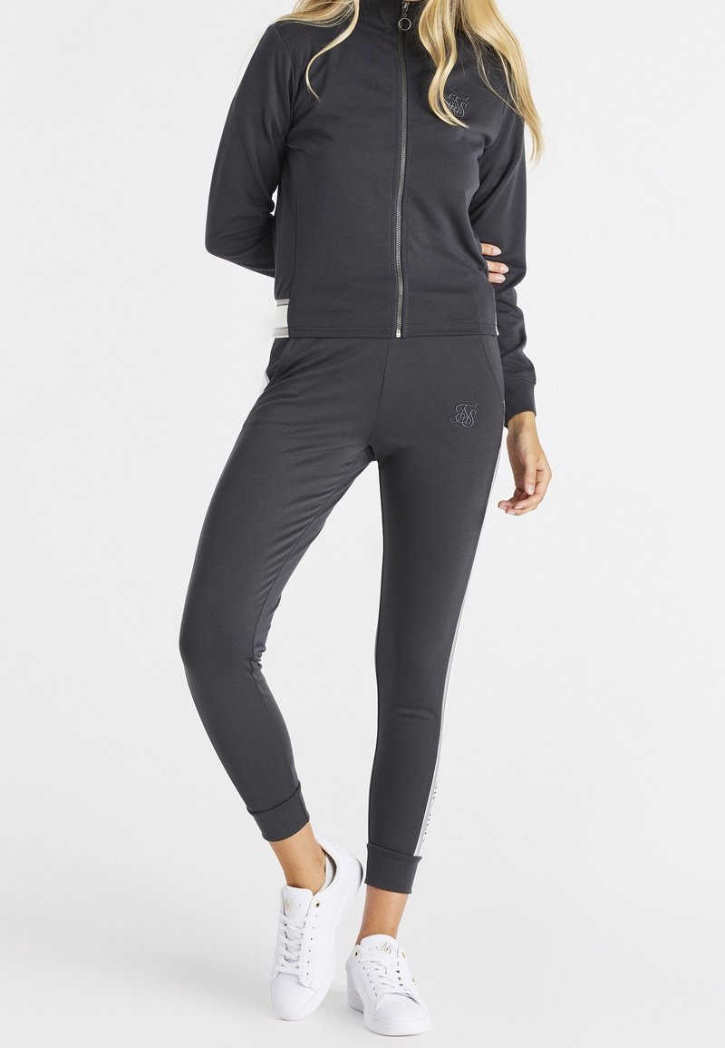 Survêtement gris foncé avec une veste zippée et un pantalon ajusté. La veste présente un col haut et un logo ; le pantalon a des rayures latérales et un détail logo.